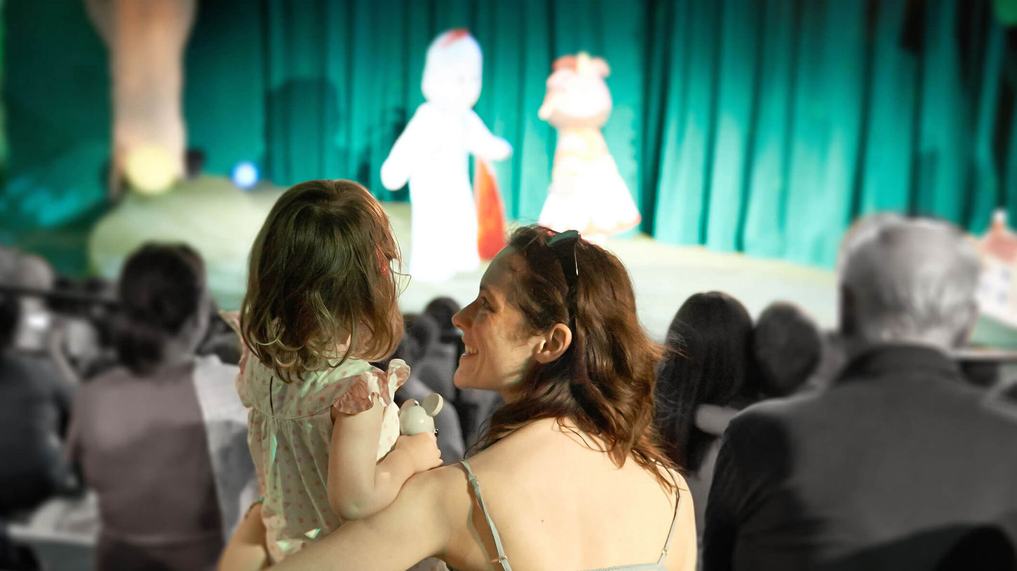 A mother and her young daughter sitting inside a theatre, with a stage in the background and people in their seats.