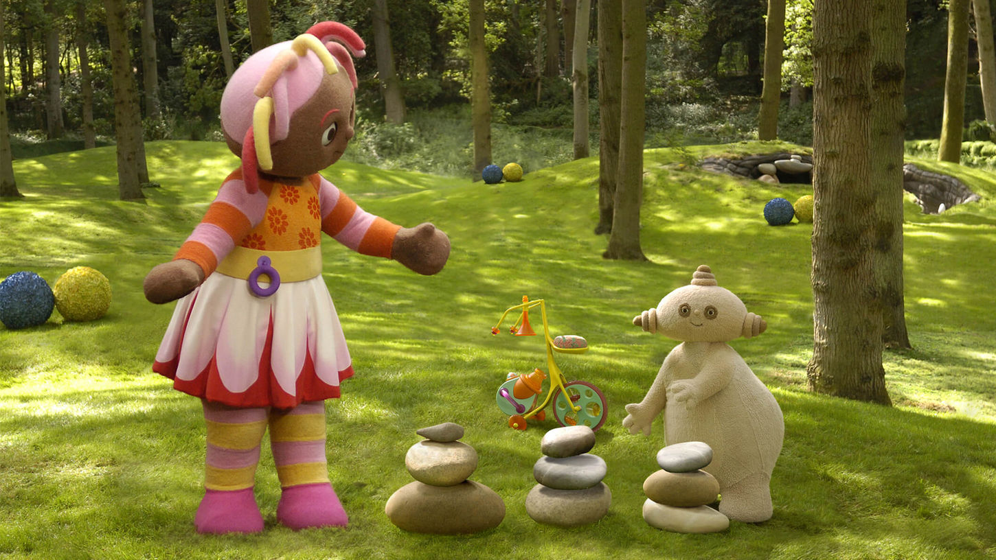 A girl with a colourful dress and hair is standing in a park beside stacked rocks and a uniquely shaped beige character.