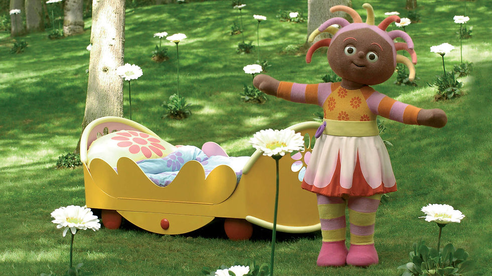 A brown, female character in a colourful dress, standing in a park with a yellow bed behind her. She is smiling and has her arms spread open.