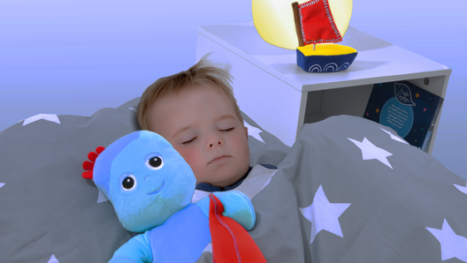A toddler sleeping in his bed beside a blue toy holding a red blanket. There is a side table with a lamp and a toy on it.