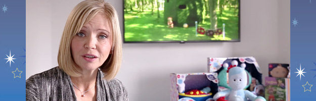 A blonde, middle-aged woman sitting in a room beside a display of toys and a TV mounted on the wall behind her.