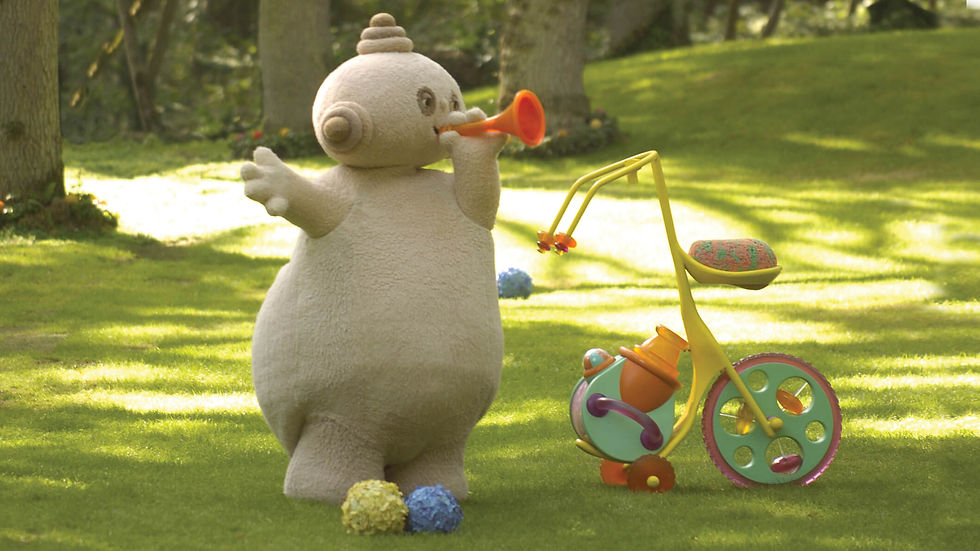 A round, beige character standing in a park, beside a tricycle and blowing through an orange horn.