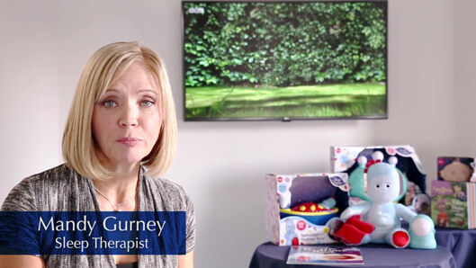 A blonde, middle-aged woman sitting in a room beside a display of toys and a TV mounted on the wall behind her. 