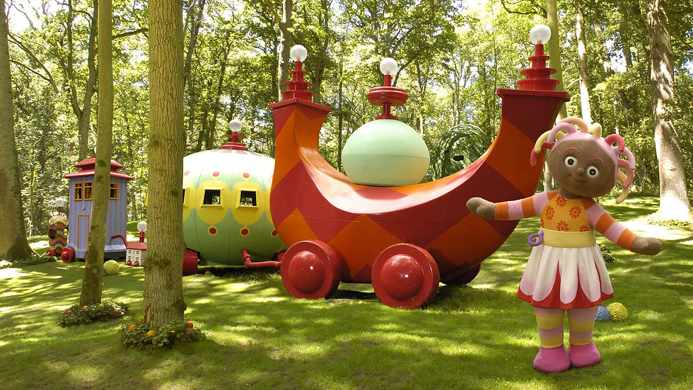 A girl with colourful, stringy hair and a colourful dress in standing on the grass in a park. Behind her is a train with three cars.