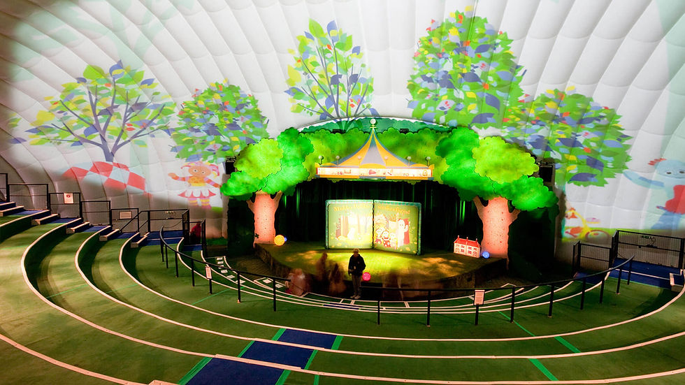 A stage inside a white tent with two trees on each side. There is a projection of colourful trees and two characters on the tent walls.