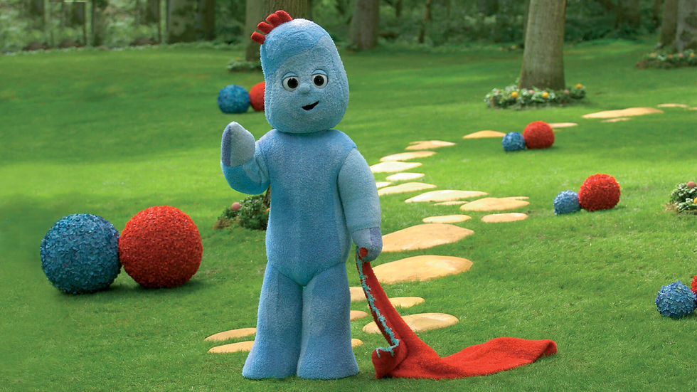 A blue character with a red patch of hair, holding a red blanket and standing in a green park with a cobble stone path in the background.