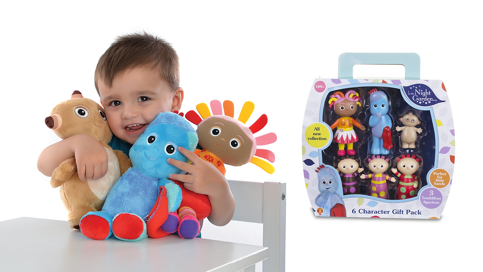 A toddler sitting at a table is smiling and hugging three soft toys. Behind the toddler is a white background and a toy packaging on the right side.